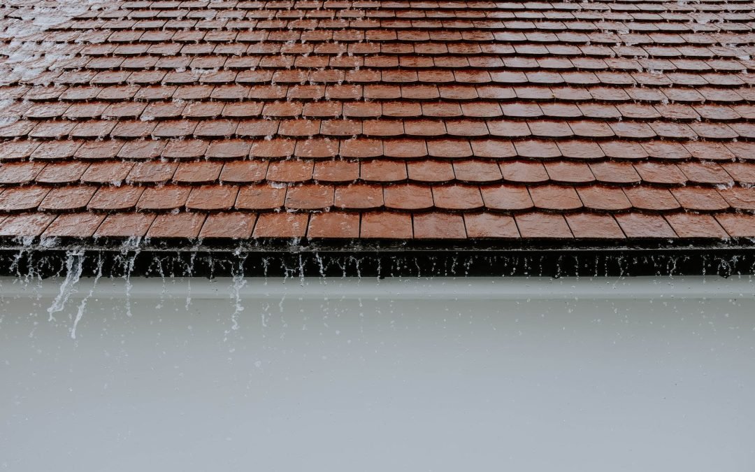 water running off a roof that has brown roof tiles and a white stucco wall beneath the roof