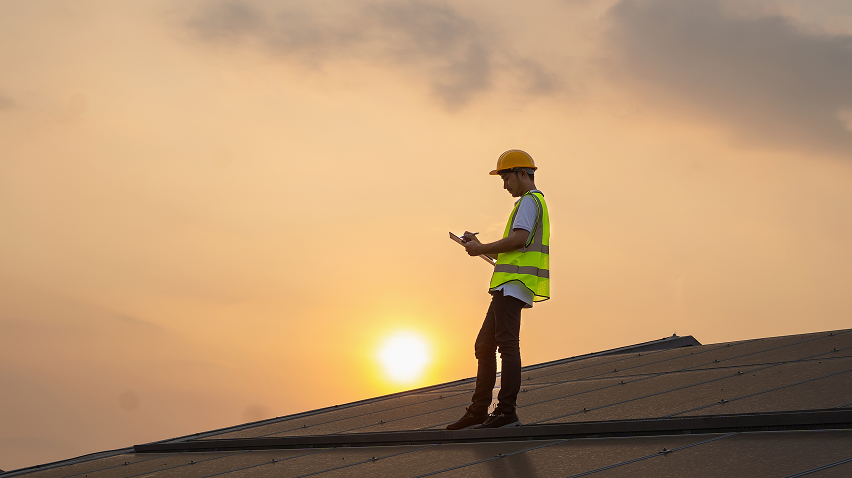Male construction worker in a hard hat and reflective vest, standing on a roof at dusk